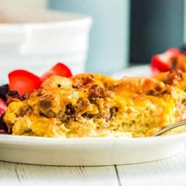 A slice of egg and sausage casserole with melted cheese on a white plate, garnished with strawberries and blueberries. A fork is placed beside the casserole.