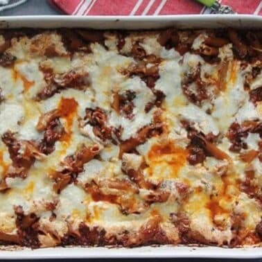 Baked pasta dish in a blue rectangular dish with melted cheese on top, placed on a gray surface with a red striped towel and a spoon beside it.