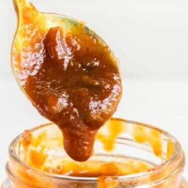 A spoon with peach barbecue sauce above an open jar of with the sauce, against a light background.