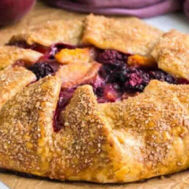 A rustic fruit galette on parchment paper, filled with peaches and blackberries, with a golden, sugared crust. Peaches and a purple cloth are partially visible in the background.