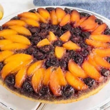A fruit upside-down cake with peach slices and blueberries on a glass serving platter, accompanied by a blue cloth and some fresh peach slices on a small plate.