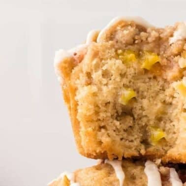 Close-up of a cut-open peach muffin with crumb topping, revealing small pieces of peach inside. Drizzles of white icing are visible on the top.