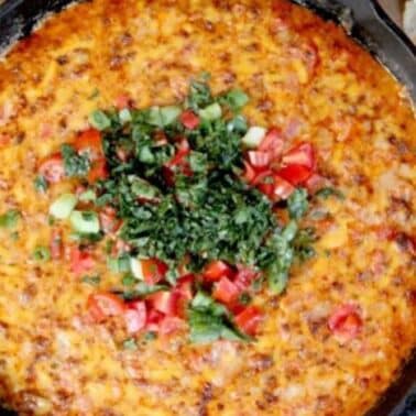 A skillet with pimento cheese queso dip with chorizo topped with diced tomatoes and cilantro, surrounded by lime wedges and tortilla chips.