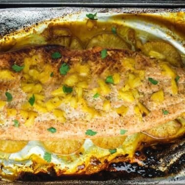 Baked salmon fillet with pineapple pieces and garnished with herbs on foil-lined baking sheet.