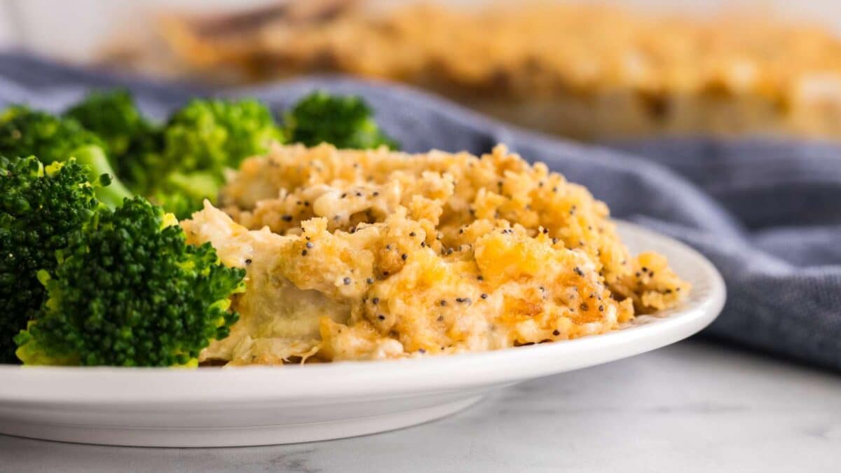 A plate of broccoli and cheesy poppy seed chicken casserole with breadcrumbs on a white surface and blue cloth.