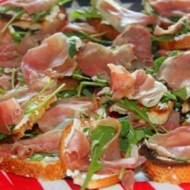 Slices of toasted bread topped with prosciutto and arugula on a red and white plate.