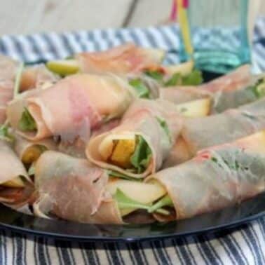 A plate of prosciutto-wrapped pears with arugula, arranged neatly on a striped cloth.