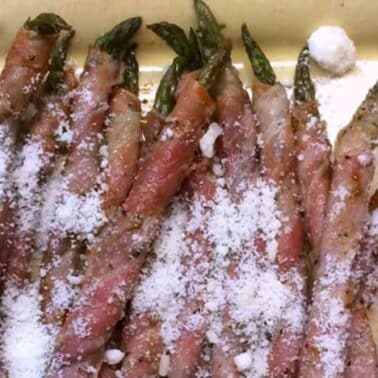 Asparagus wrapped in prosciutto, topped with grated cheese, arranged in rows on a rectangular dish.