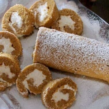 Pumpkin roll cake with cream filling, partially sliced, on a decorative platter, dusted with powdered sugar.