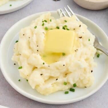 Plate of mashed potatoes topped with a pat of butter and sprinkled with chopped chives, accompanied by a fork.