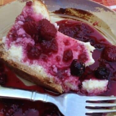 Slice of cheesecake topped with raspberry sauce on a plate, with a fork beside it. The plate sits on a plaid tablecloth.