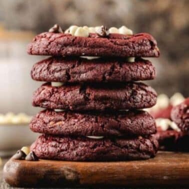 A stack of five red velvet cookies with chocolate chips on a wooden board, surrounded by more cookies and a bowl of white chocolate chips.