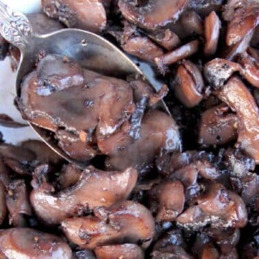 A close-up of sauteed mushrooms with a silver spoon, displaying a glossy texture and dark brown color.