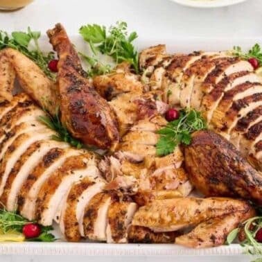 Sliced roasted turkey with herbs, lemon slices, and cranberries on a white platter.