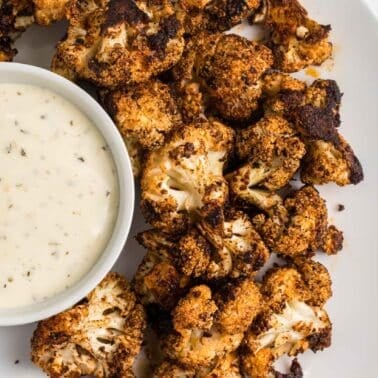 Roasted cauliflower florets seasoned with spices on a plate, accompanied by a small bowl of creamy dipping sauce.
