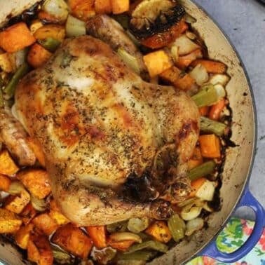 Roasted chicken in a blue pot surrounded by vegetables, including sweet potatoes, carrots, and celery, topped with herbs. A knife and a floral napkin are nearby.