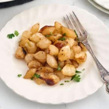 A white plate with roasted pearl onions garnished with parsley and a vintage fork on the side.