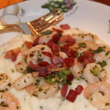 Bowl of creamy roasted poblano grits topped with shrimp, bacon pieces, and herbs. A fork rests on the edge of the bowl.
