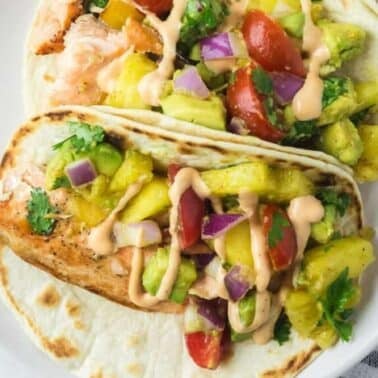Two tacos filled with salmon, diced tomatoes, onions, pineapple, cilantro, and a drizzle of sauce on a white plate.