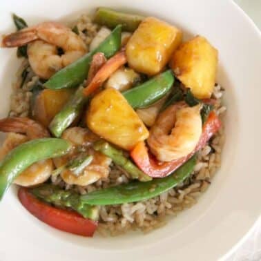 A bowl of rice topped with shrimp, pineapple chunks, snap peas, and red bell pepper slices. A fork and knife are placed adjacent to the bowl on a white surface.