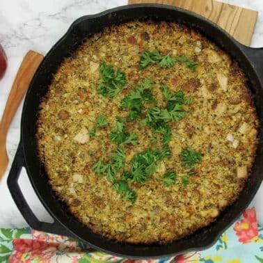 A skillet containing a dressing topped with herbs. Nearby are a wooden spatula, a red onion, fresh herbs, and a colorful cloth on a marble surface.