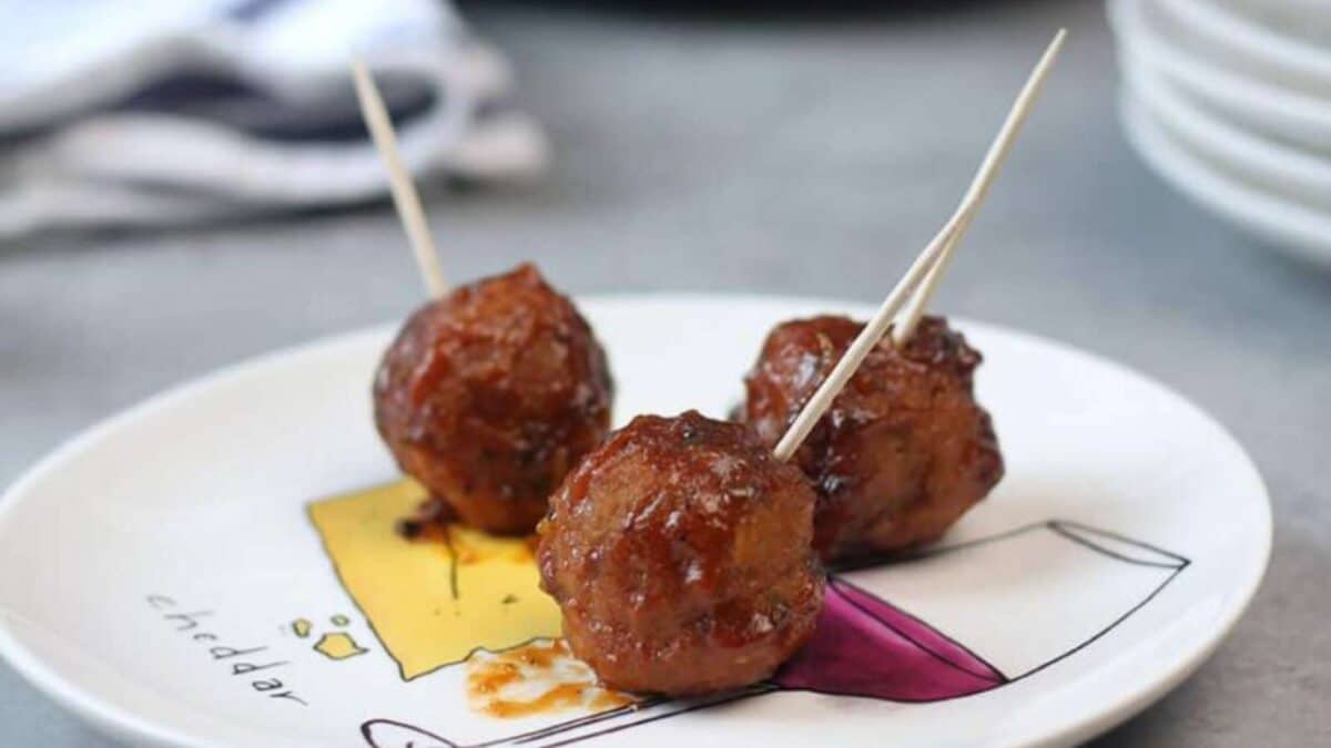 Three glazed meatballs with toothpicks are served on a white plate with a colorful design.