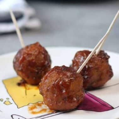 Three glazed meatballs with toothpicks are served on a white plate with a colorful design.