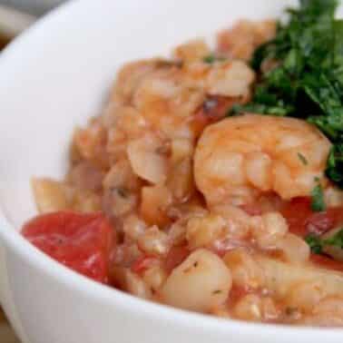 A white bowl filled with a tomato-based shrimp farro and risotto, garnished with chopped green herbs.