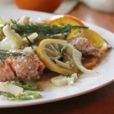 A plate with cooked salmon garnished with dill, lemon slices, fennel, and orange slices. Served on a white dish on a wooden table.