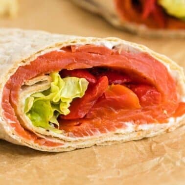Close-up of a wrap filled with smoked salmon, lettuce, and sliced red peppers, partially unwrapped on parchment paper.
