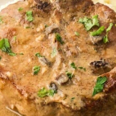 A cooked pork chop with mushroom gravy, garnished with parsley, served alongside mashed potatoes.