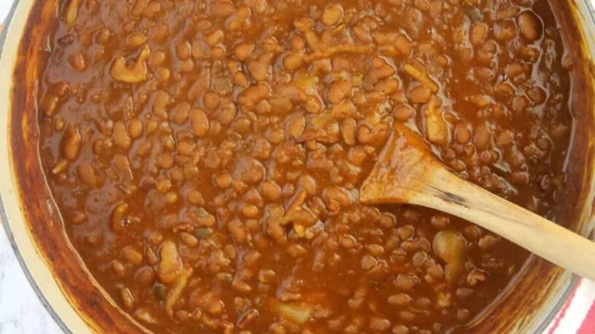 A pot filled with baked beans in a thick sauce, with a wooden spoon stirring them.