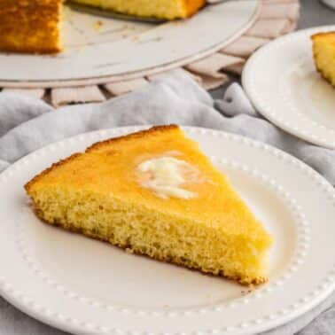 A slice of cornbread with a pat of butter on a white plate. Another slice and forks are in the background on a grey tablecloth.