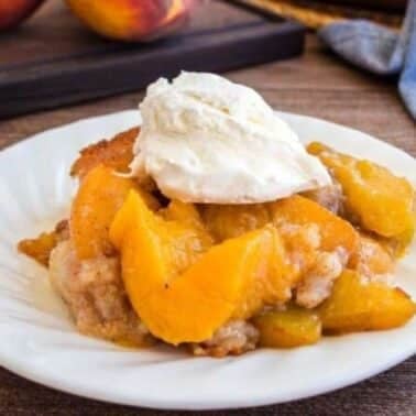 Peach cobbler with a scoop of vanilla ice cream on a white plate, placed on a wooden table.