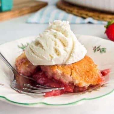 Slice of strawberry cobbler topped with a scoop of vanilla ice cream on a decorative plate, with a fork beside it. Fresh strawberries are in the background.