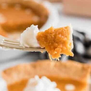 A fork holds a piece of sweet potato pie topped with whipped cream. In the background is a whole pie and a checkered cloth.