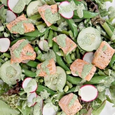 A salad with mixed greens, cucumber slices, radishes, snap peas, and chunks of salmon, drizzled with a green dressing on a white plate.