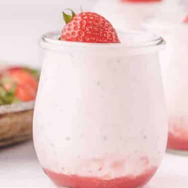 A glass jar of strawberry mousse topped with a fresh strawberry. Other jars and strawberries are visible in the background.