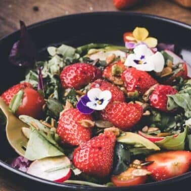 A salad with strawberries, mixed greens, cherry tomatoes, seeds, and edible flowers in a black bowl, accompanied by a gold spoon.