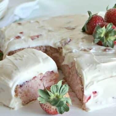 A sliced strawberry cake topped with white frosting and strawberries is displayed on a table.