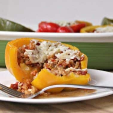 A yellow bell pepper stuffed with rice and meat topped with cheese on a white plate, beside a fork. A dish of green and red stuffed peppers is in the background.