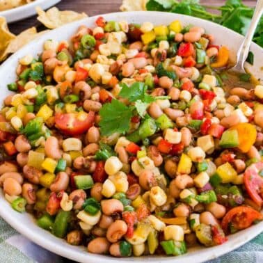 A bowl of colorful bean salad with tomatoes, corn, bell peppers, and a cilantro garnish, placed on a green and white cloth. Tortilla chips and fresh cilantro are in the background.