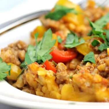Close-up of a plate with a ground beef dish, topped with chopped parsley and diced vegetables, including red peppers. A fork is partially visible beside the dish.