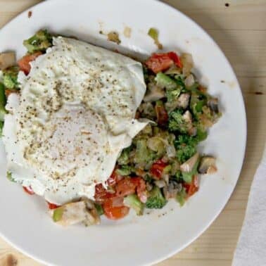 A plate with fried egg on top of a mixed vegetable hash, featuring broccoli, tomatoes, and mushrooms, on a wooden table next to a fork and a napkin.