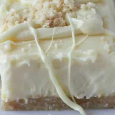 Close-up of a white chocolate cheesecake bar with a crumbly crust and a drizzle of white chocolate on top.