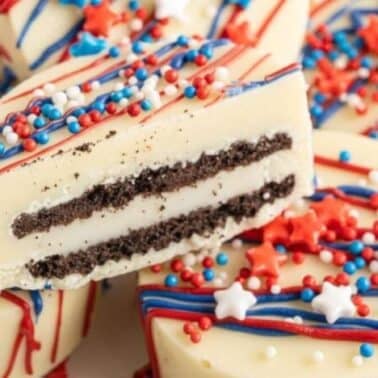 White chocolate covered Oreos with red, white, and blue decorations are stacked, with one cookie showing a cross-section of chocolate layers inside.