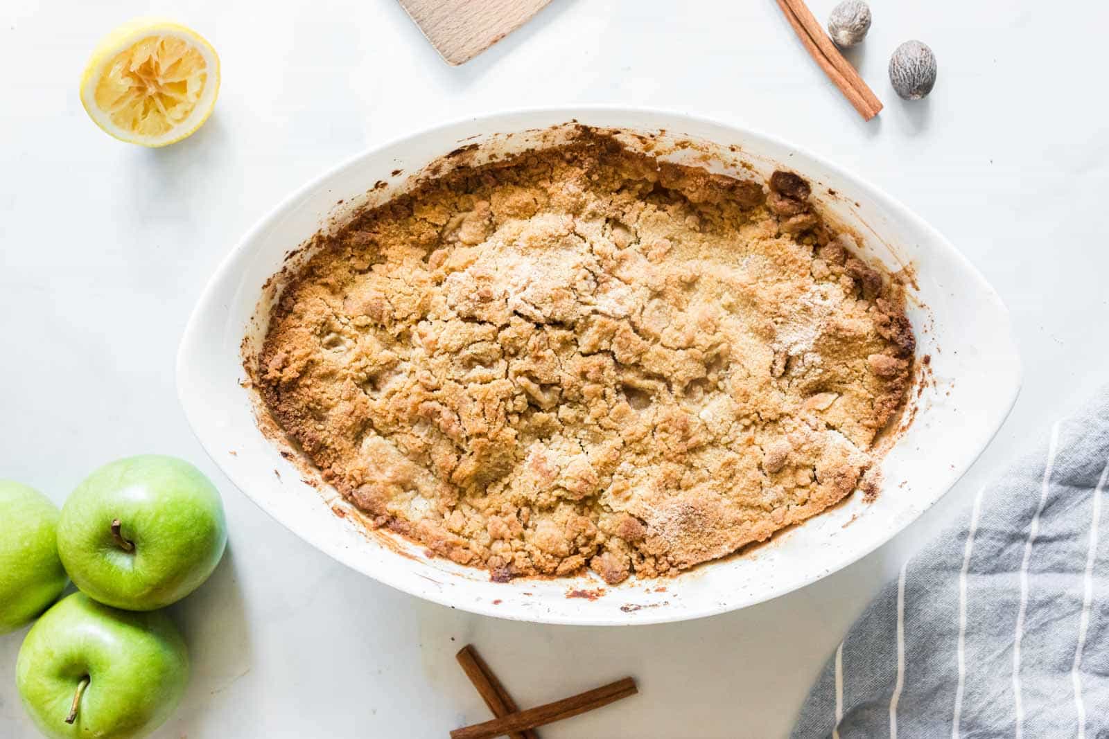 A baked apple crisp in an oval dish on a white surface, surrounded by green apples, a lemon half, cinnamon sticks, a grater, and whole nutmeg.