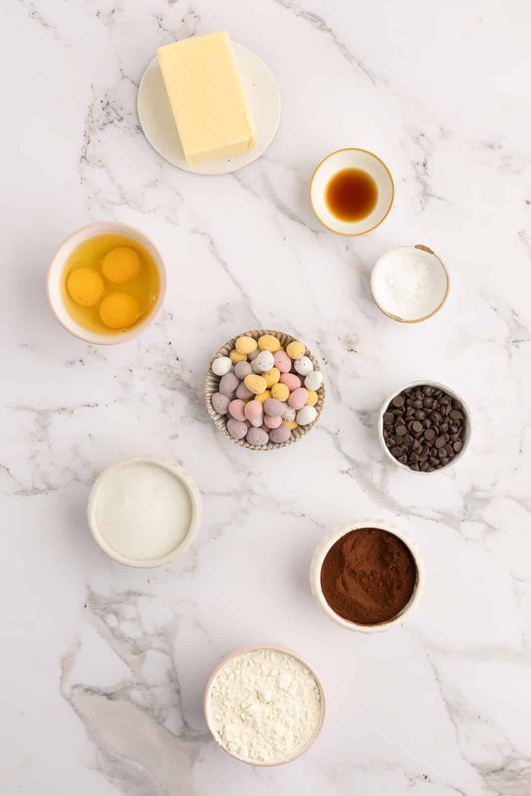 Various baking ingredients including eggs, butter, vanilla, sugar, chocolate chips, cocoa, flour, baking powder, and mini chocolate eggs arranged on a marble surface.