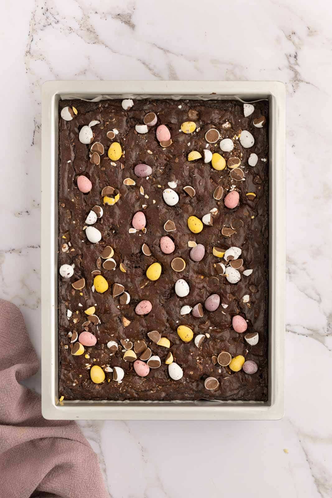 A baking pan filled with chocolate brownies topped with colorful candy eggs on a marble countertop. A light pink cloth rests beside the pan.