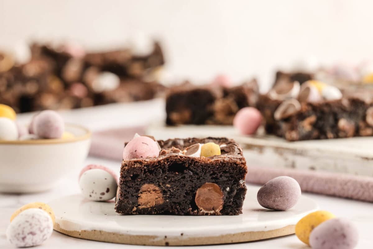 A chocolate Easter brownie square on a white plate, adorned with colorful candy eggs, surrounded by more candies and a bowl in the background.